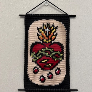 May include: Crocheted wall hanging featuring a Sacred Heart design. The heart is red, surrounded by a green thorn crown and flames. The background is beige, framed by a black border. The piece is suspended by a black cord and rods.