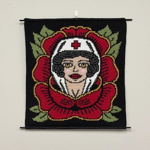 May include: A black crochet wall hanging featuring a nurse's face within a red rose. The nurse wears a white cap with a red cross. The rose has green leaves and a yellow outline. The artwork is mounted on black rods.