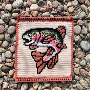 May include: A square, crocheted wall hanging with a rainbow trout design. The fish is detailed with pink, green, and orange against a cream background. The border and hanging loop are orange. The piece is suitable for a home.