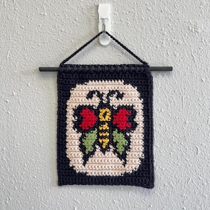 May include: A handmade, square wall hanging featuring a butterfly design. The butterfly is embroidered with red, yellow, and green details, set against a cream background and a black border. The piece is suspended from a black rod.