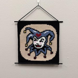 May include: A handmade, square wall hanging featuring a jester design. The jester has a smiling face, a blue hat with a red heart, and is set against a beige background. The piece is framed in black and suspended by a black rod and string.