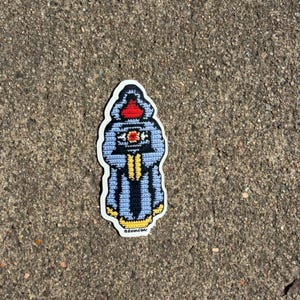 May include: A sticker featuring a blue, yellow, and red crochet design of a hooded figure. The figure has a red hood, a yellow and black design on the chest, and a blue robe. The sticker has a white border and the text "@dixonfiber".