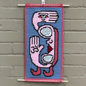 May include: A handmade crochet wall hanging featuring a whimsical pink figure against a blue background. The figure has a hand, eyes, and a mouth, outlined in pink, with a pink border. The artwork is displayed with wooden dowels.