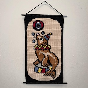 May include: A handmade, crocheted wall hanging featuring a brown seal balancing balls and wearing a circus hat. The seal is set against a beige background with a black border. The wall hanging is suspended by a black rod and string.