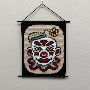 May include: A handmade, crocheted wall hanging featuring a clown face. The clown has a white face with red accents, yellow eyes, and a blue and yellow hat. The artwork is framed in black and hangs from a black rod.