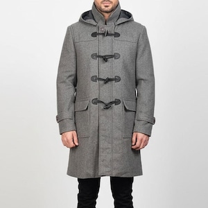 Könnte beinhalten: Ein grauer Woll-Dufflecoat mit Kapuze und schwarzen Knebelverschlüssen. Der Mantel hat zwei Vordertaschen und eine klassische, lange Silhouette. Der Mantel ist ein vielseitiges Oberbekleidungsstück.