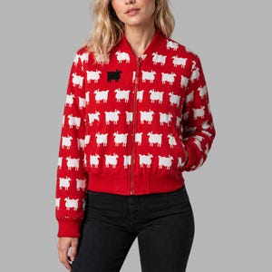 Pode incluir: Casaco bomber vermelho com um padrão de ovelhas brancas e uma ovelha preta no peito. O casaco tem fecho de correr, punhos e bainha canelados vermelhos. É feito de um material macio e confortável.
