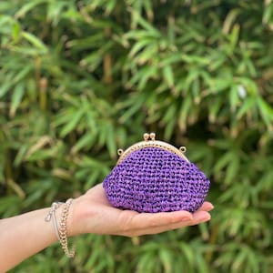 Könnte beinhalten: Eine kleine, lila gewebte Clutch mit einem goldfarbenen Verschluss. Die Clutch wird in einer Hand vor einem Hintergrund aus grünem Laub gehalten. Die gewebte Textur der Clutch ist sichtbar, und der Verschluss hat ein dekoratives, abgerundetes Design.