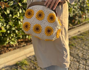 May include: Cream-colored crochet handbag with a brown leather strap and clasp. The bag features a pattern of yellow and brown sunflowers. The bag is held by a person wearing a tan trench coat and white sneakers.