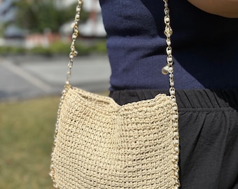 Könnte beinhalten: Beige gehäkelte Umhängetasche mit goldener Kette. Die Tasche besteht aus gewebtem Material und ist mit kleinen weißen Perlen verziert. Der Riemen ist mit goldenen Kettengliedern, kleinen weißen Perlen und einigen lila Edelsteinen verziert.