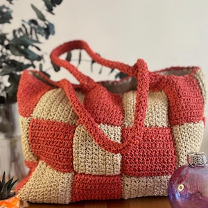 May include: A large, crocheted tote bag with a checkered pattern in beige and coral. The bag has two thick, crocheted handles.