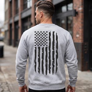 May include: A light gray sweatshirt with a distressed black American flag design on the back. The flag features black stars and stripes, creating a patriotic theme. The sweatshirt is a crew neck style.