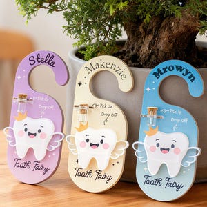 Op de afbeelding: Drie kleurrijke tandfee deurhangers met de namen Stella, Makenzie en Mrowyn. Elke hanger heeft een lachende tand met vleugels en de tekst "Tooth Fairy". Ze hebben een klein flesje met een kurk en de woorden "Pick Up" en "Drop Off".