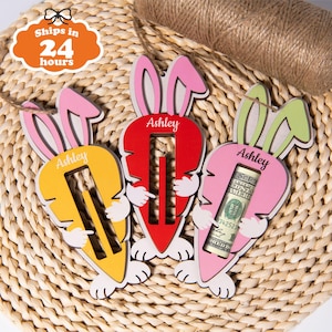 Könnte beinhalten: Drei hölzerne Osterhasen-Karotten-Geldhalter in Gelb, Rot und Rosa. Jeder hat einen Schlitz für Bargeld und den Namen "Ashley" aufgedruckt. Die rosa Karotte enthält eine aufgerollte Banknote. Die Hasenohren sind rosa und grün.