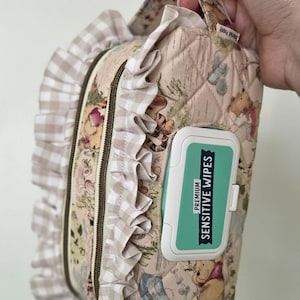 Pode incluir: Uma bolsa de cosméticos acolchoada com um padrão de Winnie the Pooh, com acabamento em babados e fecho de correr. Inclui um recipiente verde-azulado e branco de "Sensitive Wipes".