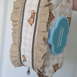 May include: A beige and white quilted baby wipes container with a teddy bear pattern. The container has a ruffled edge and a blue plastic dispenser that reads "THE LARGEST BABY WIPES".