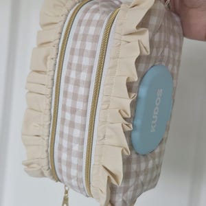 May include: A beige and white gingham patterned baby wipe case with a light blue lid that reads "KUDOS". The case has a beige ruffle trim and a gold zipper. It has a handle for carrying.