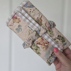 Peut inclure: Un article en tissu matelassé avec un fond beige orné d'une illustration fantaisiste. L'article a une bordure vichy et est orné de petits nœuds en tissu. Le texte "Emerald Poppy" est visible.