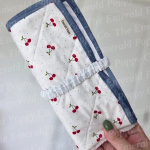 Könnte beinhalten: Weißes, gestepptes Stoffetui mit Kirschmuster und blauem Jeansbesatz. Das Etui wird mit einem weißen Gummiband verschlossen. Der Text "Emerald Poppy" ist auf dem Etui sichtbar.