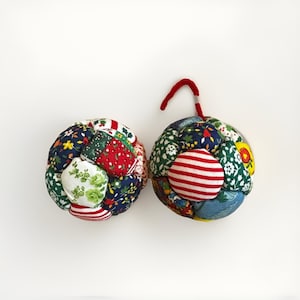 Peut inclure: Deux ornements sphériques en tissu fabriqués à partir de divers patchs matelassés de coton coloré. Les motifs incluent des imprimés floraux, des rayures rouges et blanches et des feuillages verts. Un ornement possède une boucle en tissu rouge.