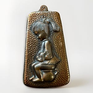 Puede incluir: Placa de metal de tono bronce vintage con un relieve detallado de un niño pequeño sentado en un orinal, sosteniendo un objeto pequeño. La placa tiene un fondo texturizado y un lazo en la parte superior para colgar. El niño lleva un vestido y tiene una coleta.