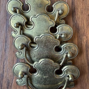 Puede incluir: Un juego de cuatro tiradores de cajón de latón con una placa decorativa festoneada y un acabado envejecido. Cada tirador incluye un asa curva oscilante unida a dos postes, mostrando una forma ornamentada con arañazos superficiales visibles.