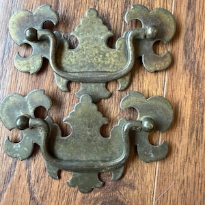 Puede incluir: Dos tiradores de cajón de latón a juego con una placa decorativa que presenta un remate central puntiagudo y elementos laterales curvos. Cada tirador tiene un asa basculante unida a dos puntos de montaje circulares con pátina.