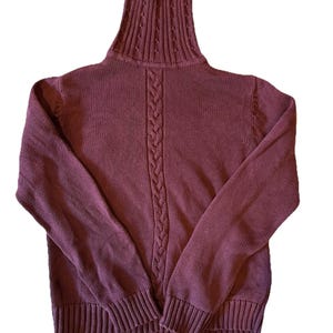 May include: A burgundy turtleneck jumper with a cable knit design down the centre and ribbed cuffs. The high collar has a textured pattern. The long sleeves extend to ribbed cuffs. The jumper is made of a soft, knitted material.