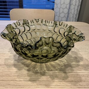 Puede incluir: Un cuenco de vidrio verde vintage con un borde con volantes. El cuenco tiene un patrón texturizado en el exterior y un borde festoneado. El cuenco está sobre una mesa de madera de color claro.