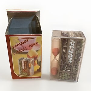 May include: A vintage "Ecology 3 Minute Timer" with a box featuring a teacup and egg illustration. The timer includes an hourglass and a clear container filled with various beans. The box is red and yellow.