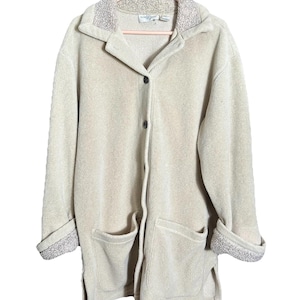 May include: A beige fleece jacket with a collared neckline, two front pockets, and button closures. The cuffs and collar lining feature a textured, light gray fabric. The jacket is hanging on a pink hanger.