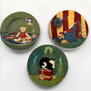 May include: Three round ceramic plates featuring Christmas illustrations. One plate shows a teddy bear with a toy train, another depicts Santa Claus in a chair, and the third features a black and white cat sitting on a red gift box with a green wreath.