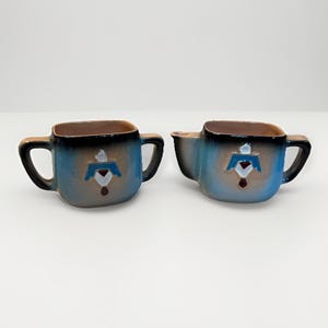 Peut inclure: Deux tasses en céramique de forme carrée avec une anse. Les tasses sont bleues avec un bord marron et un motif décoratif sur le devant. Le motif est un oiseau stylisé ou une forme géométrique avec des accents rouges et blancs.
