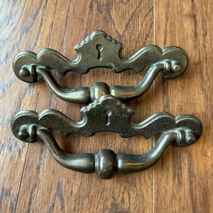 Peut inclure: Deux poignées de tiroir en laiton de style ancien avec une patine sombre et vieillie. Chaque pièce présente une plaque décorative avec une découpe centrale en forme de trou de serrure et une poignée mobile incurvée fixée aux deux extrémités.