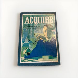 May include: A vintage Acquire board game box featuring a blue and green illustration of a person holding glasses. The text reads ACQUIRE and HIGH ADVENTURE IN THE WORLD OF HIGH FINANCE. The design includes stylized city buildings and a desk.
