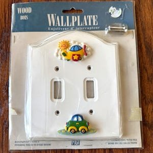 May include: A white double light switch wallplate in original packaging. The plate features a yellow airplane with a red propeller and a smiling sun above, and a green car below. Text on the packaging includes WOOD BOIS and WALLPLATE.