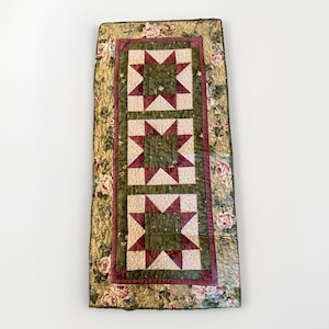 May include: A rectangular quilted table runner featuring three star patterns in the center. The stars are made of cream and burgundy fabric, set against a green background. The border has a floral pattern in shades of pink, green, and cream.