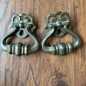 May include: Two matching brass drawer pulls featuring a decorative backplate with a scrollwork top and a hinged, teardrop-shaped handle. The metal has a muted, aged finish with visible patina, showcasing a symmetrical design and sturdy construction.