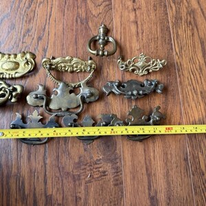 May include: A collection of assorted brass drawer pulls and furniture handles in various shapes and sizes, displayed with a yellow measuring tape showing measurements in inches and centimeters to indicate their scale and dimensions.
