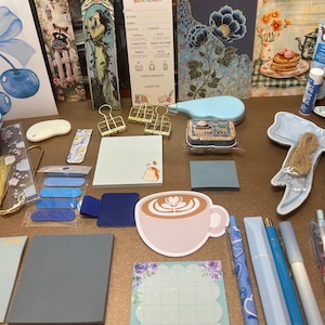 May include: A collection of stationery items, including bookmarks, sticky notes, pens, and paper clips. The items are in shades of blue, pink, and gold, with decorative floral and coffee cup designs. There is a book review card and a small cupcake box.