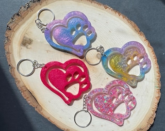 May include: Four heart-shaped keychains with paw prints. The keychains are in shades of pink, red, blue, and purple. Each keychain has a silver keyring attached. The keychains are on a wooden surface.