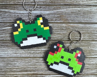 May include: Two pixel art frog keychains. The frogs are green with black outlines and white bellies. One frog has yellow accents, the other has red accents. Each keychain has a silver ring and chain.