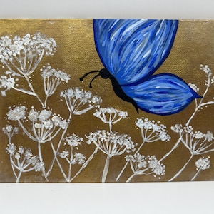 Op de afbeelding: Een blauwe vlinder met witte vleugels vliegt over een veld met witte bloemen op een gouden achtergrond.