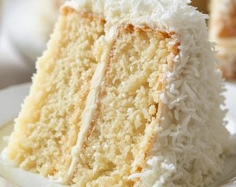 May include: A slice of layered cake with white frosting and shredded coconut on a white plate with a gold rim. The cake has a light yellow sponge and a creamy filling. Another cake is visible in the background.