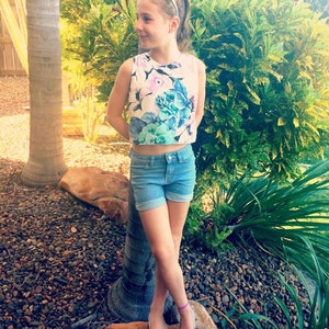 May include: A young person wearing a white crop top with a floral print and blue denim shorts. They are standing on a rock with a palm tree in the background. The person is wearing silver shoes and a silver headband.