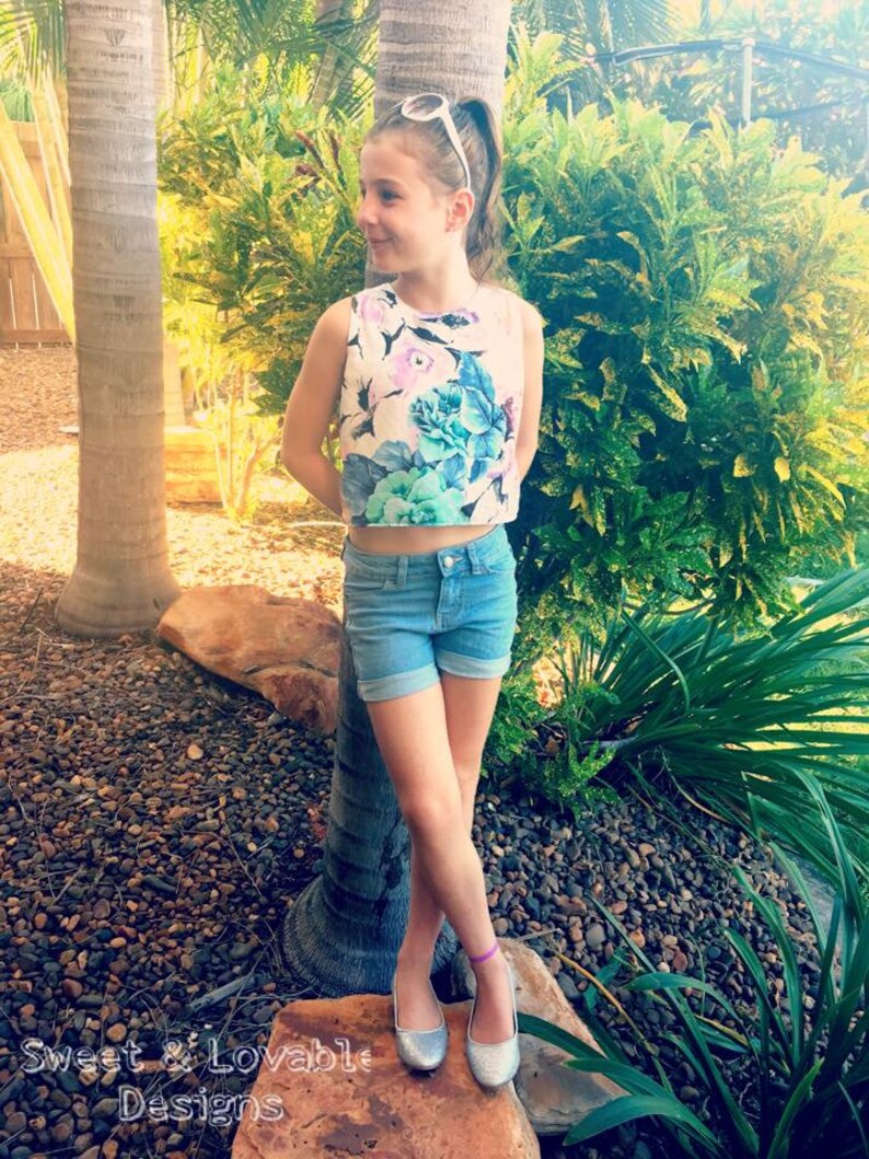 May include: A young person wearing a white crop top with a floral print and blue denim shorts. They are standing on a rock with a palm tree in the background. The person is wearing silver shoes and a silver headband.