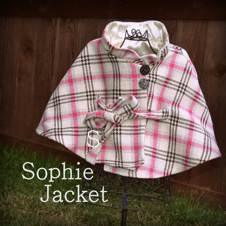 Puede incluir: Una capa de cuadros blancos y rosas con ribete negro y un bot&oacute;n negro. La capa est&aacute; sobre un soporte negro y el texto "Sophie Jacket" est&aacute; debajo de la capa.