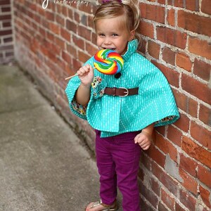 Puede incluir: Una ni&ntilde;a peque&ntilde;a con una capa turquesa con un cintur&oacute;n marr&oacute;n y pantalones morados sostiene una piruleta colorida. Est&aacute; de pie frente a una pared de ladrillo.