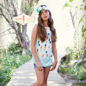 Puede incluir: Una persona joven lleva una blusa azul claro sin mangas con un estampado abstracto colorido, pantalones cortos de mezclilla y sandalias gladiadoras marrones. Lleva una corona de flores con flores blancas y azules. La persona está de pie en una pasarela de madera en un entorno boscoso.