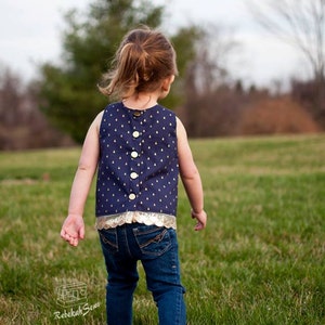May include: A young person wearing a navy blue sleeveless top with gold accents and a gold button closure. The top has a white and gold pattern. The person is wearing blue jeans and is walking barefoot on green grass.